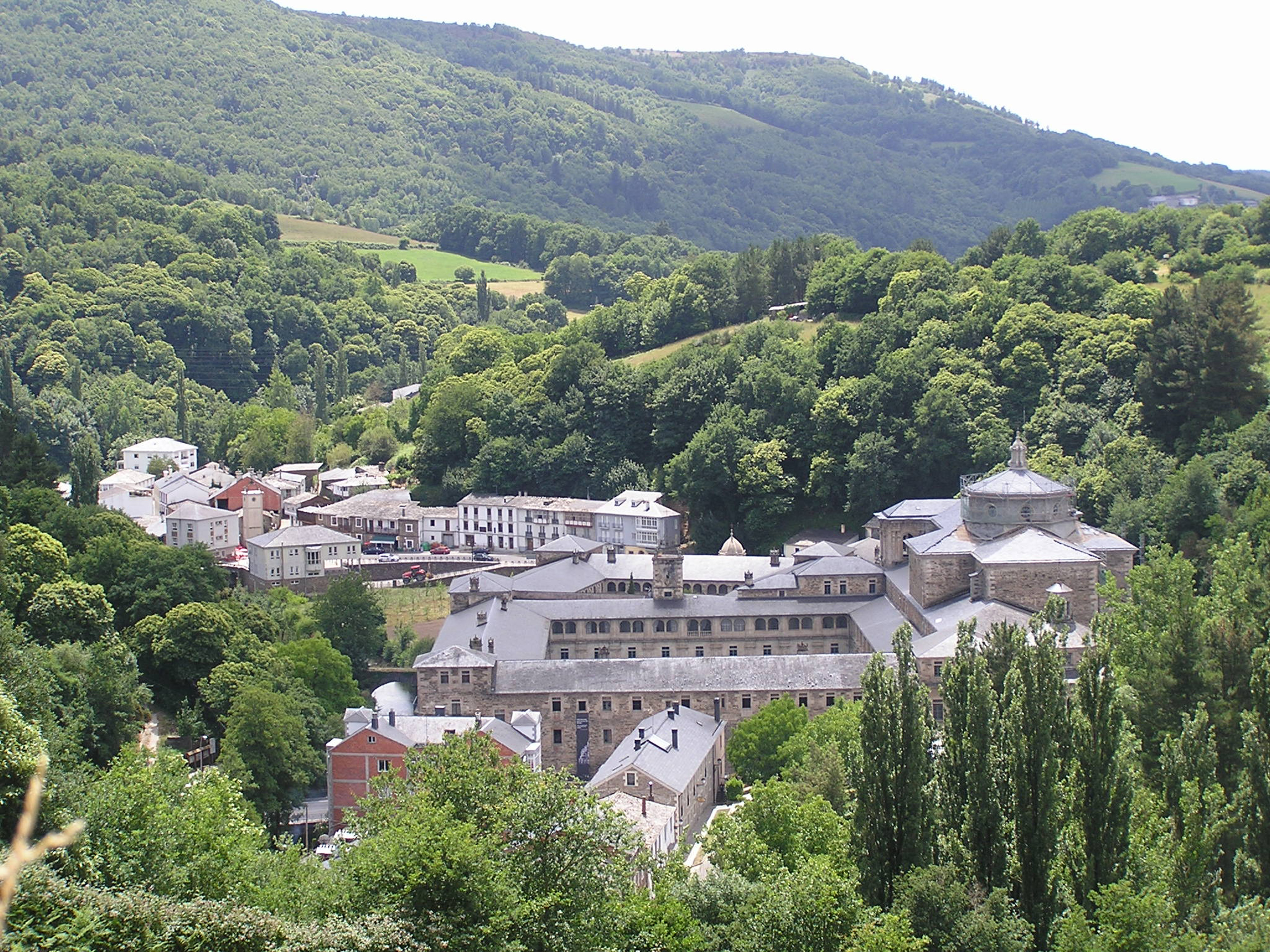 A color photograph of the monastery of San Julián de Samos. The site is seen at a distance and form slightly above, as though the photographer is on a hill above the site. A group of buildings of various layout and construction are seen in a small valley between green forested hills.