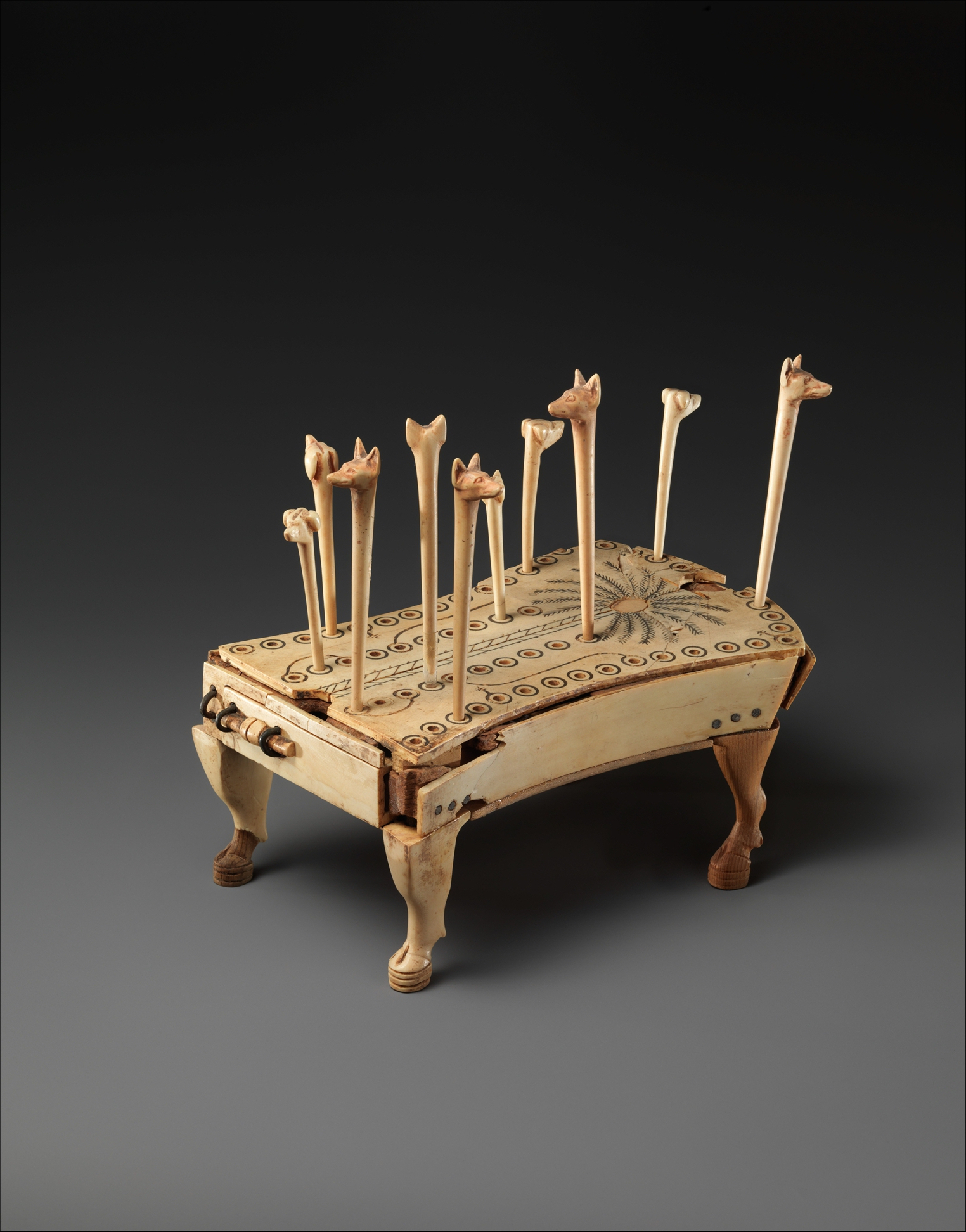 A small ivory game board with holes for spaces and pegs with the heads of dogs and jackals inserted into them.